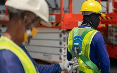 Two construction workers wearing safety gear and helmets; one is fastening a safety harness to the other.