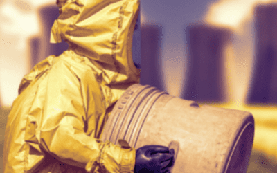 Person in a yellow hazmat suit and black gloves handling a large metal barrel with industrial cooling towers in the background.