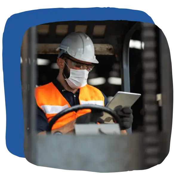 A worker wearing a hard hat, face mask, and orange safety vest operates a tablet while sitting in industrial equipment.
