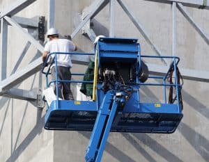 A construction worker wearing a helmet stands on a blue aerial lift, working on a steel structure attached to a concrete wall.