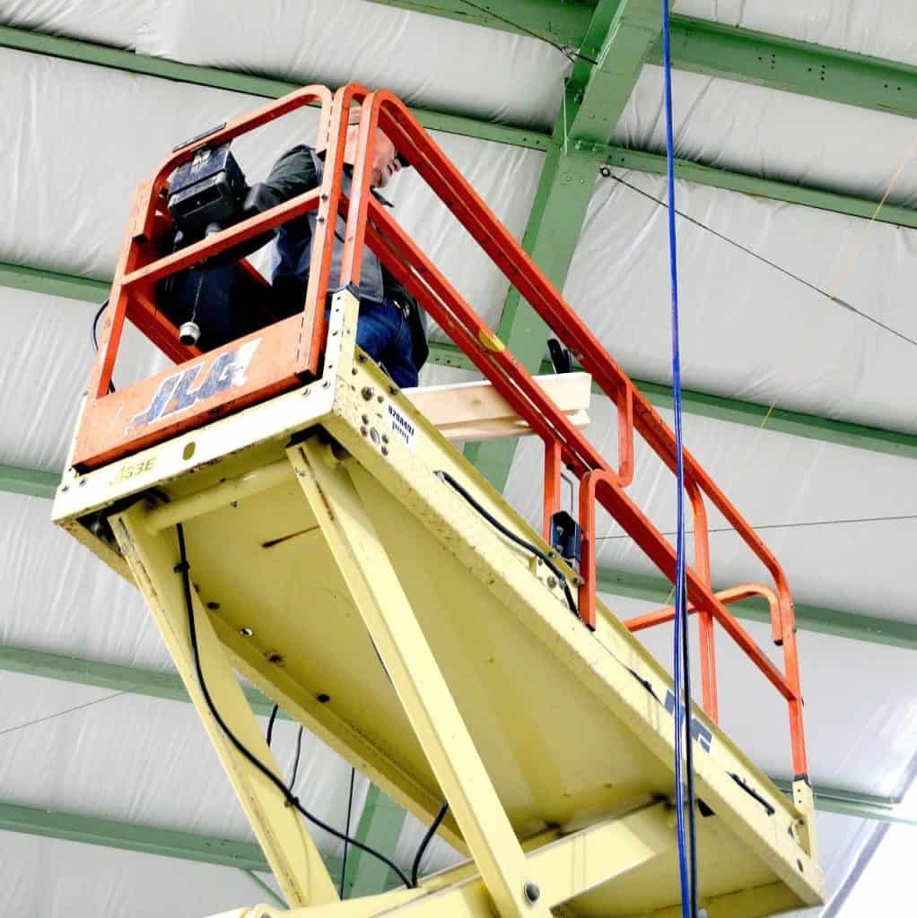 A person operates a scissor lift inside a building with a metal frame and insulated ceiling panels.
