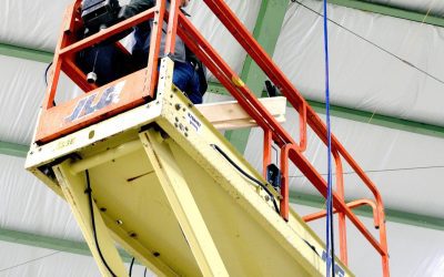 A person operates a scissor lift inside a building with a metal frame and insulated ceiling panels.